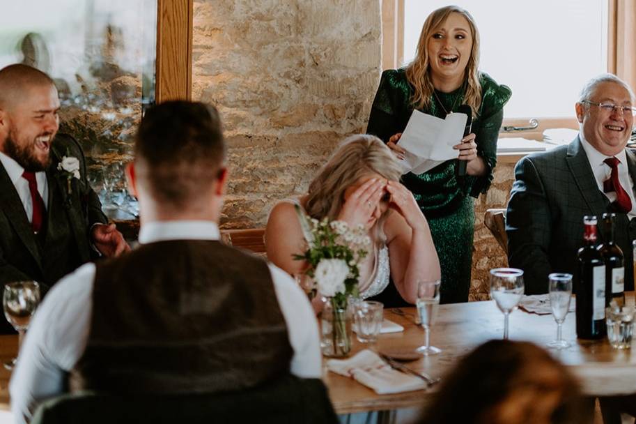 a maid of honour wearing a green metallic dress stands at the top table laughing as she delivers funny wedding readings in a speech with the couple and guests laughing too