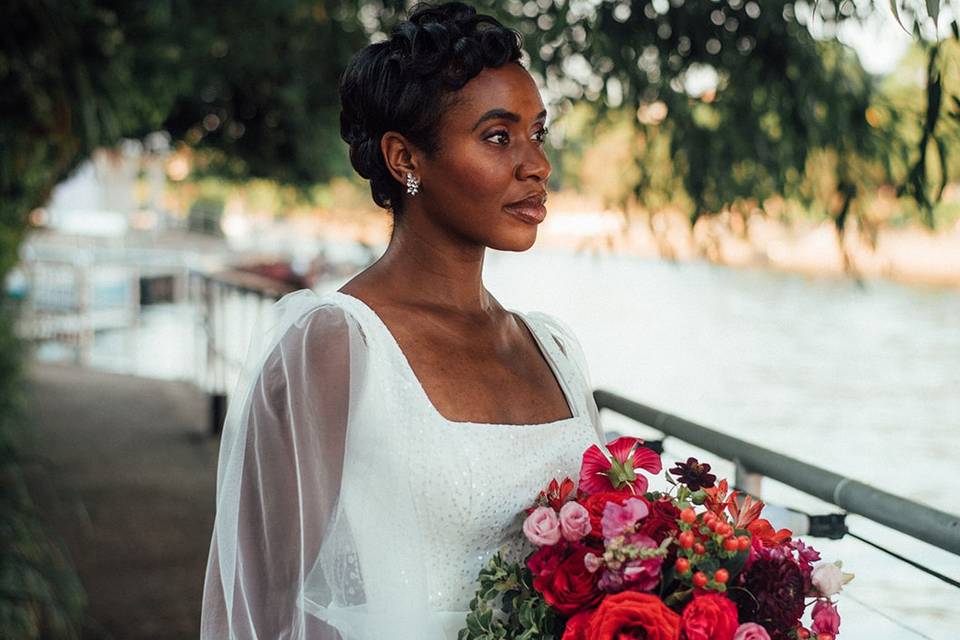 Black bride with elegant wedding makeup holding a berry toned bouquet looking out over a river