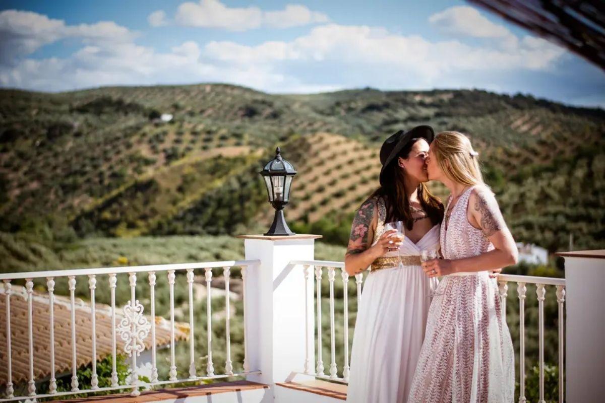 two brides in beautiful wedding dresses kiss as they hold champagne on a balcony with rolling hills in the background