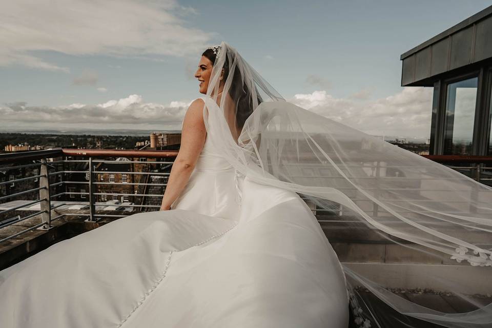 A smile bride with her back to the camera on a roof, as her white dress and veil billows in the wind