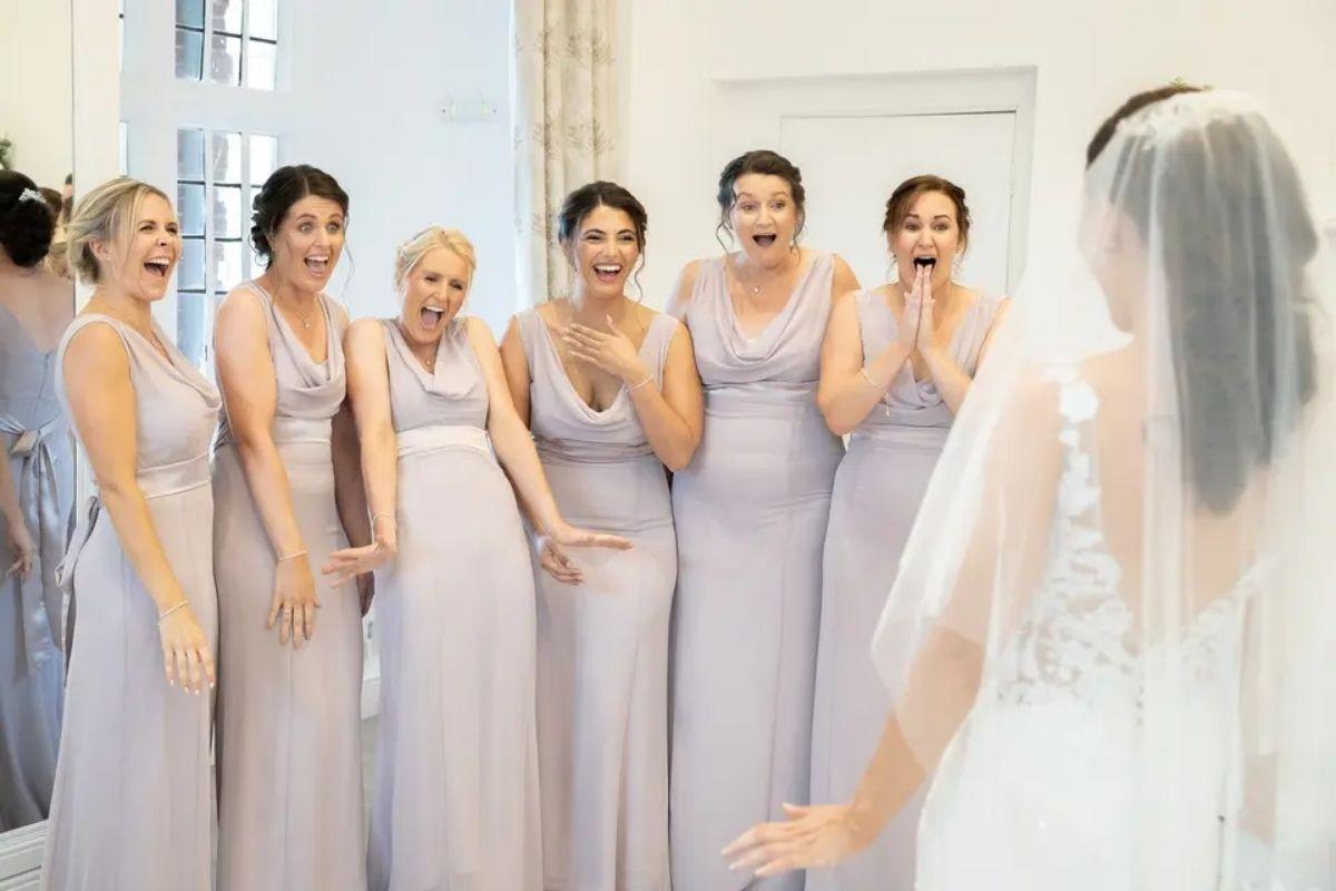 six bridesmaids in lavender coloured bridesmaid dresses gasp and look emotional as they see the bride in her wedding dress for the first time