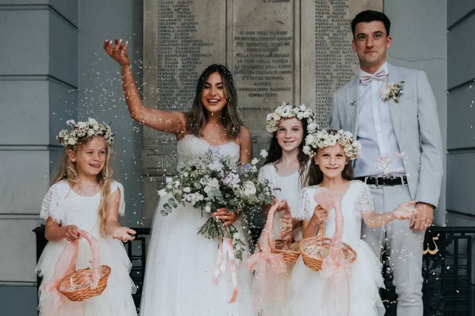 A bride, groom and two flower girls with wedding flower bouquets and crowns standing outside their venue throwing confetti