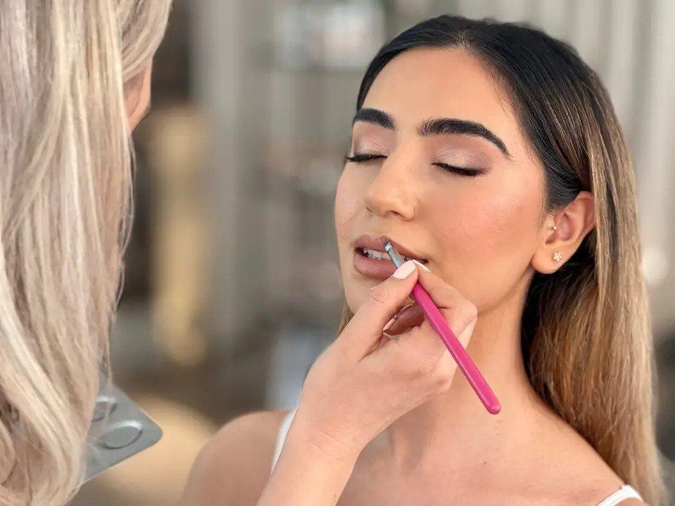 A bride is seated with her eyes closed while an MUA applies her lipstick