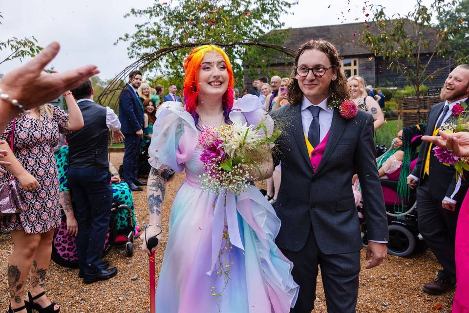 eliza wears a rainbow pastel wedding dress and uses a walking stick to walk past guests with michael at their wedding