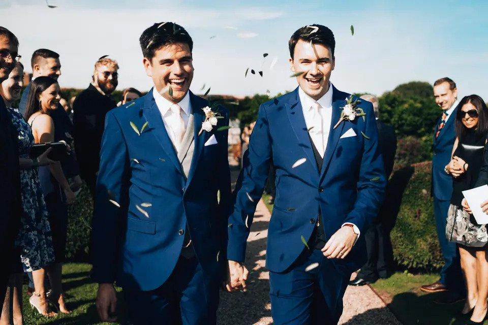 Two grooms walk hand in hand outdoors while their guests throw confetti