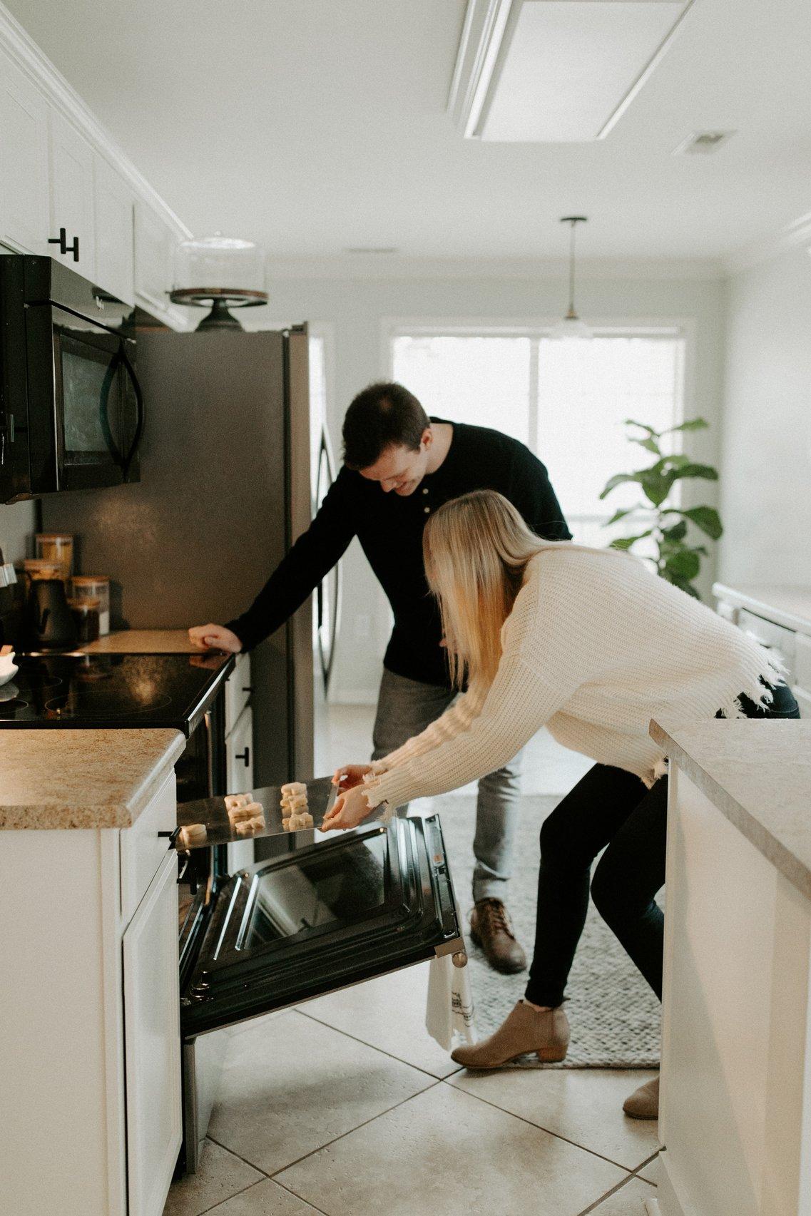 A woman and a man laughing in the kitchen as they put biscuits into the oven