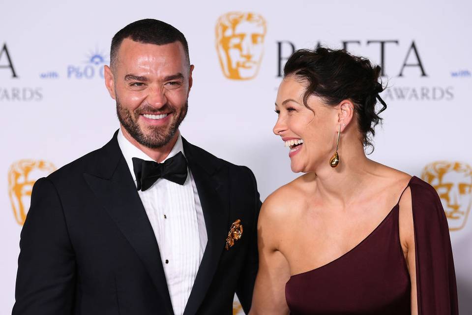 Emma and Matt Willis standing together and laughing in front of a Bafta 2024 backdrop for the red carpet, each dressed in black tie