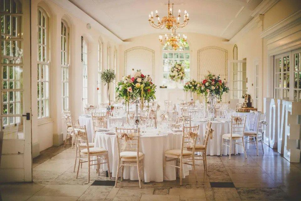 Conservatory room set for wedding meal with white round tables, Chiavari chairs, floral centrepieces and gold chandeliers