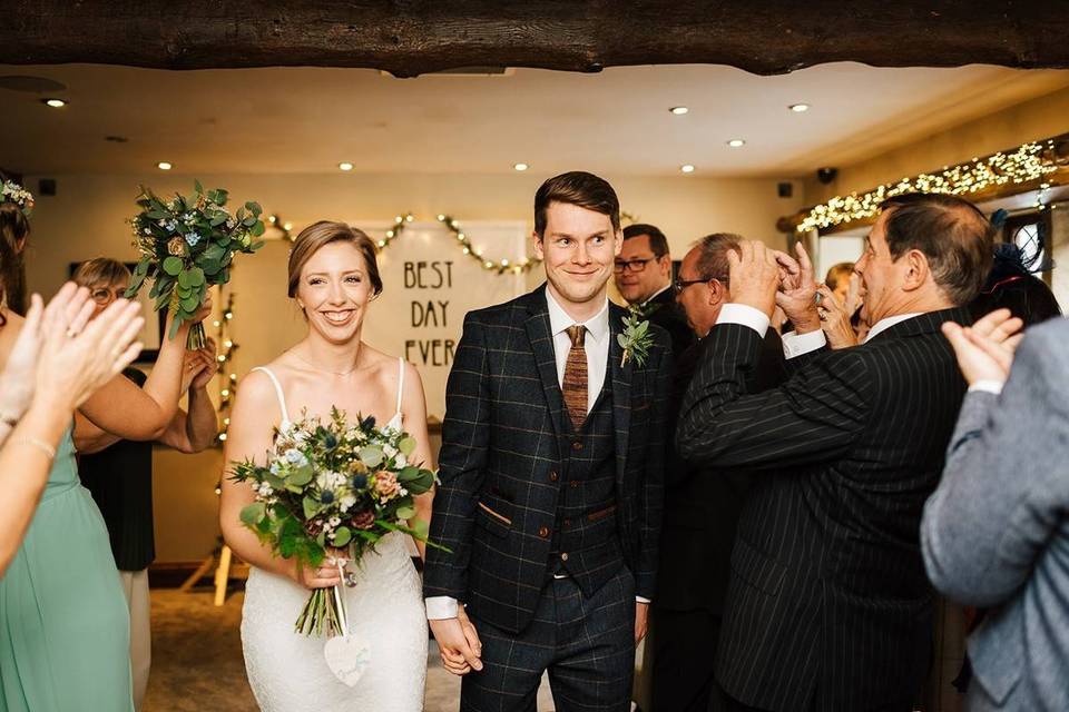 Groom in a navy check suit holding the hand of his bride, who carries a large spring bouquet, as they walk down the aisle into a crowd of applauding guests in a rustic wedding venue