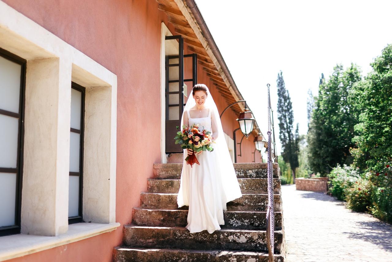 bride in a veil and dress walking out of a pink house on a sunny day holding a pink bouquet