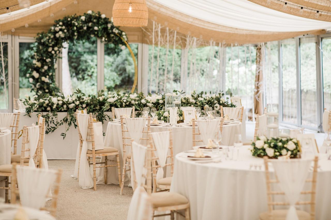Interior of a glass walled marquee style structure decorated for a wedding with lots of white florals and greenery