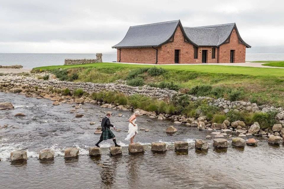 Bride and groom step across stones on a lake leading into the sea beside a boathouse venue