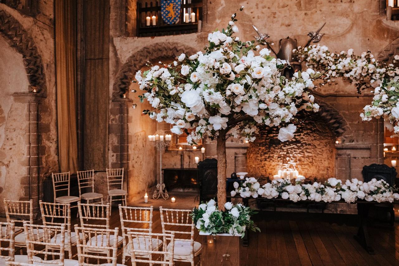 A grand room of a castle lit by candlelight and filled with white flowers, cream chairs, and a stone archway dressed for a wedding