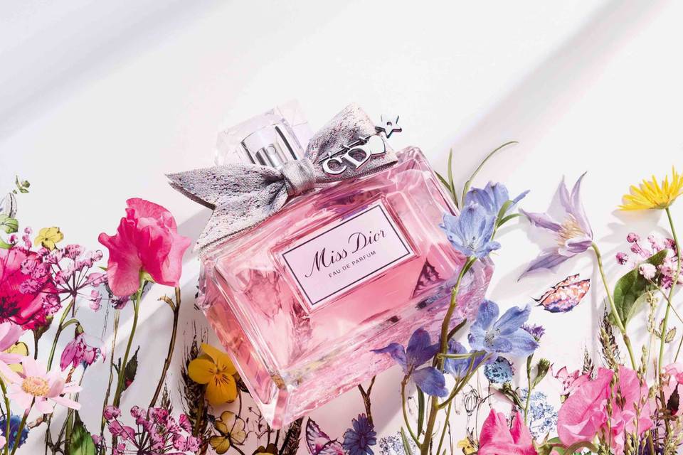 a rectangular bottle of wedding perfume Miss Dior with a silver bow on it against a white background with flowers coming up from the bottom