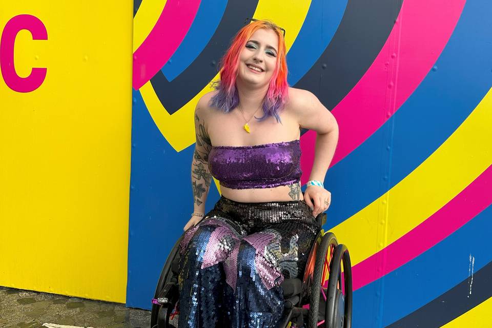 Disabled activist Eliza Rain posing in their wheelchair in front of a colourful wall, whilst wearing a sequinned crop top and trousers