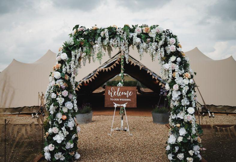 A large floral wedding arch and wooden rustic welcome sign in front of a festival style tipi wedding venue