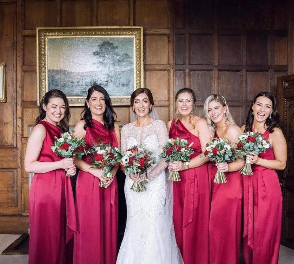 A bride and her bridesmaids dressed in red dresses pose for the camera in front of a wood panelled wall with a gold framed picture