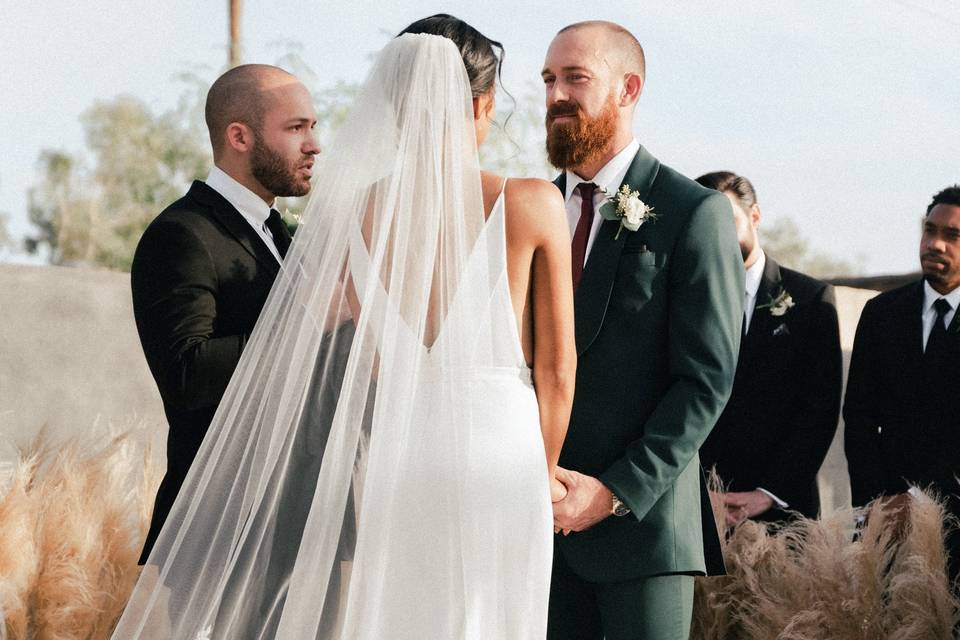 a bride and groom holding hands in front of their celebrant in an outdoor wedding with the best men standing to the side