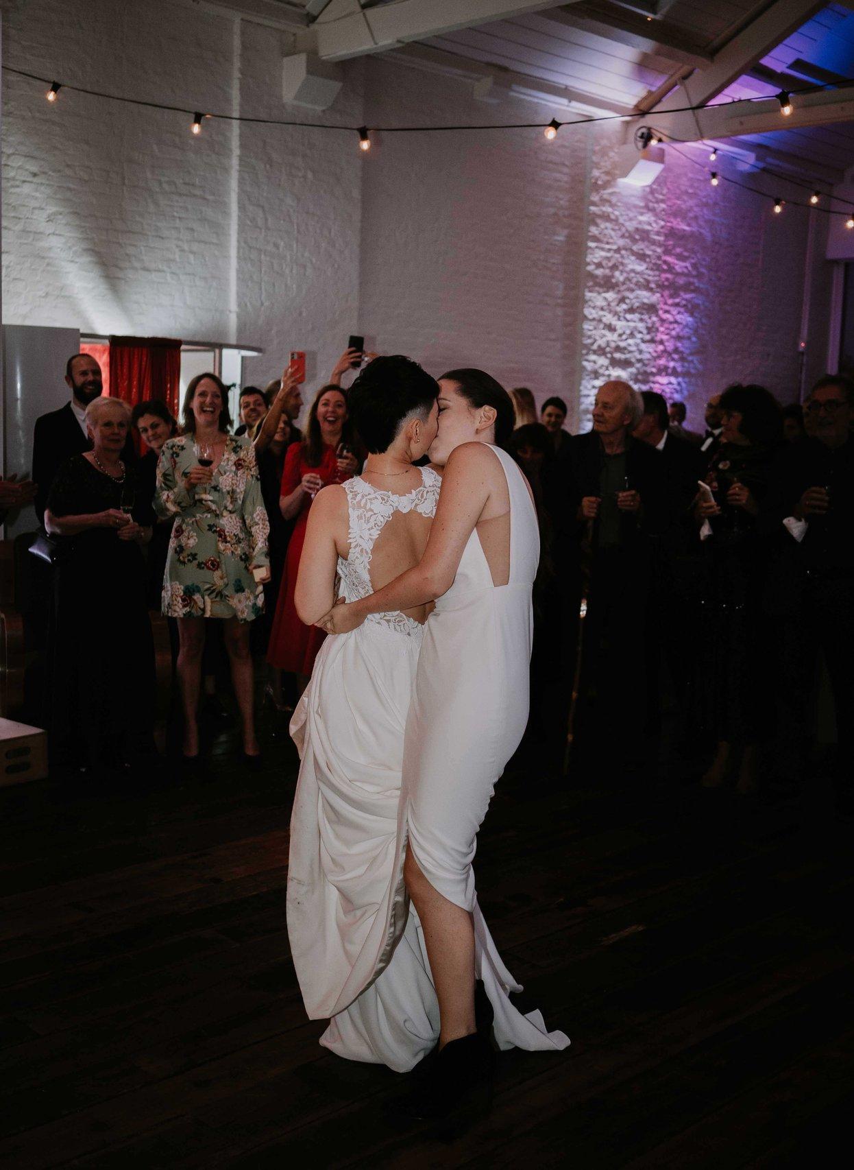 The brides kiss on the dance floor as guests watch on