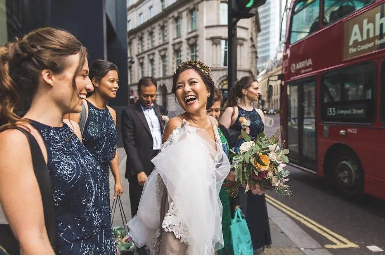 Smiling bride laughing with her bridesmaids - her hair is in an updo with a gold floral headband and her cheeks have a dewy glow