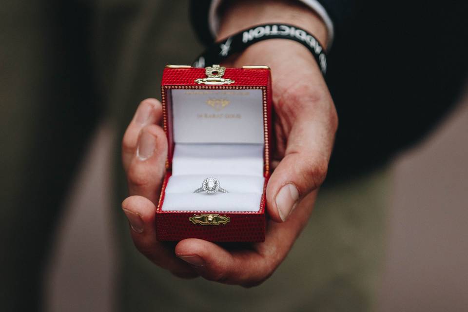 White man's hand wearing a black and white wristband holding a red ring box with a white gold and diamond oval halo ring inside