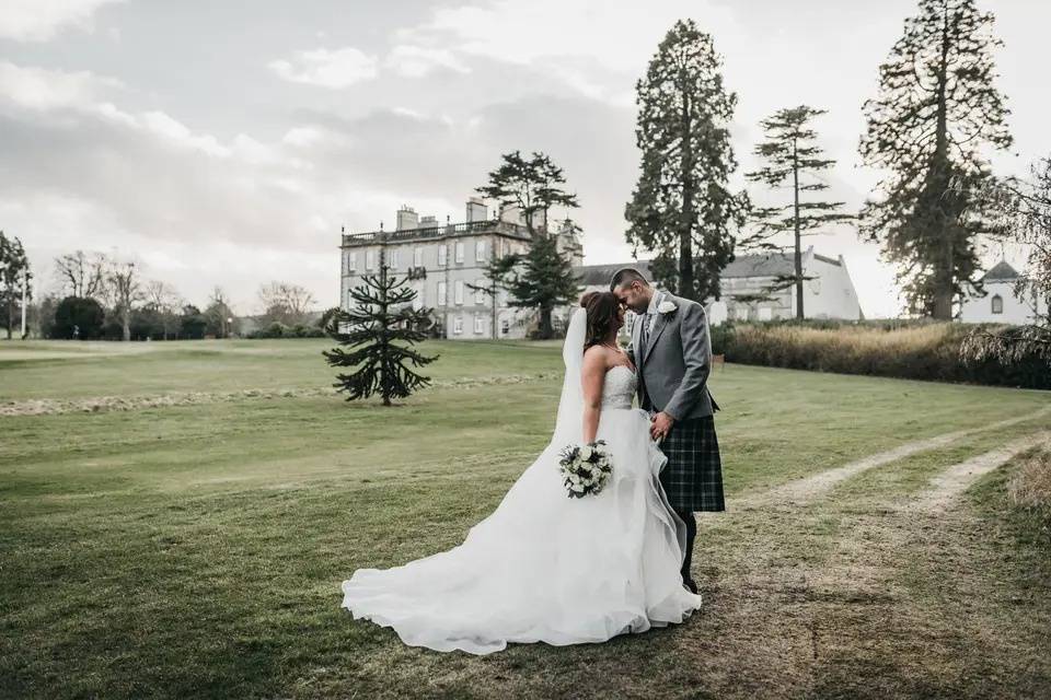 Newlyweds embrace on a manicured lawn with a gorgeous Edinburgh wedding venue in the background