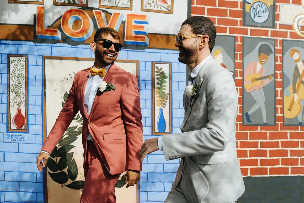 Two grooms in pastel toned suits walking together and smiling outside of a wedding venue in front of a painted wall