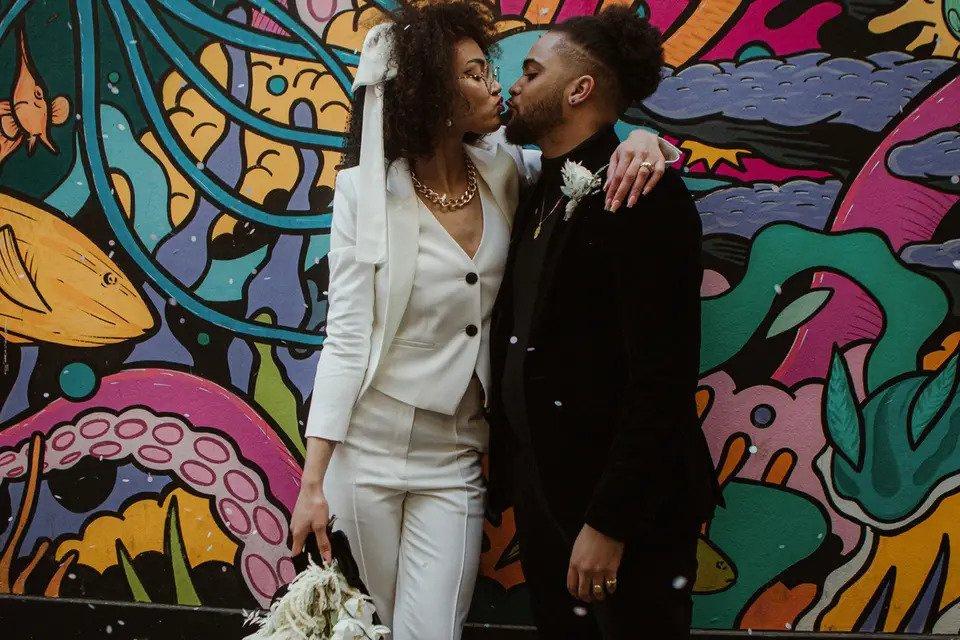 Mariée en costume blanc embrassant un marié en costume noir devant un mur de graffitis