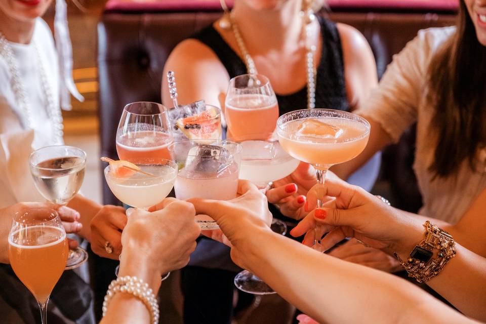 Group of white women clinking glasses containing pink and pale white cocktails