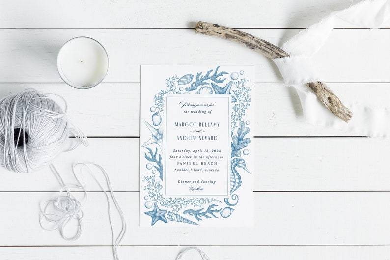 A white beach themed wedding invitation with blue shell border on a white wooden surface