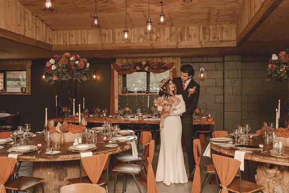 Bride and groom embrace in a tiled room with wood panelled roof, round wooden tables, chairs, dinner candles and floral centrepieces