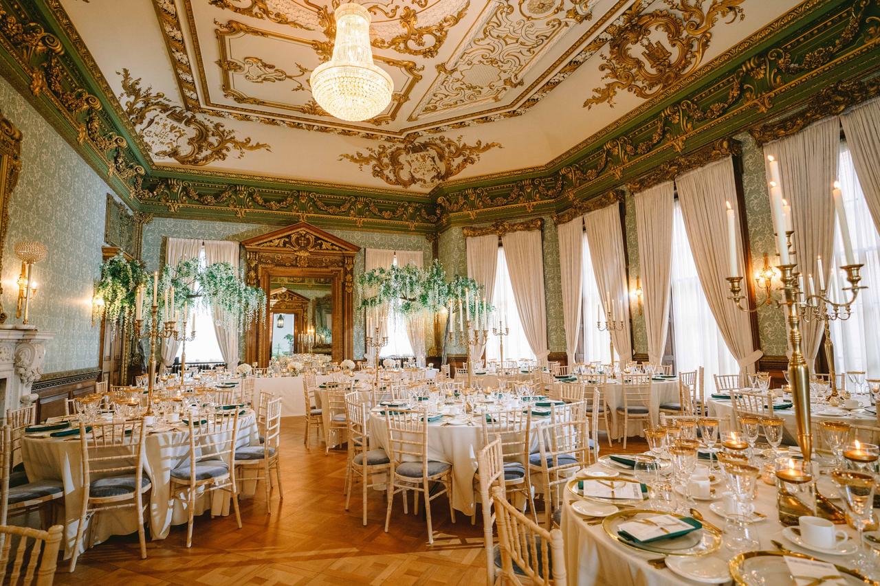Interior of a ballroom with ornate ceiling set up for a wedding reception with gold decor and round tables.