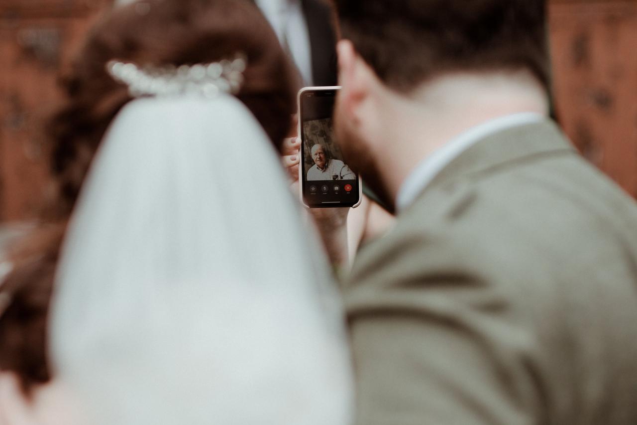 Backwards shot of a bride and groom FaceTiming a white man in a shirt on a smartphone