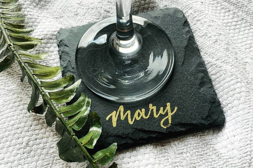 Black slate coaster with 'Mary' in gold metallic writing, with a sprig of fern and a wine glass