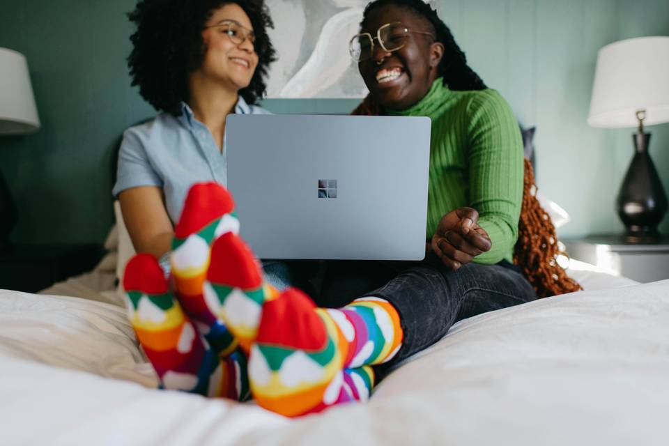 Two women on the bed wearing colourful socks and laughing at something on a laptop screen