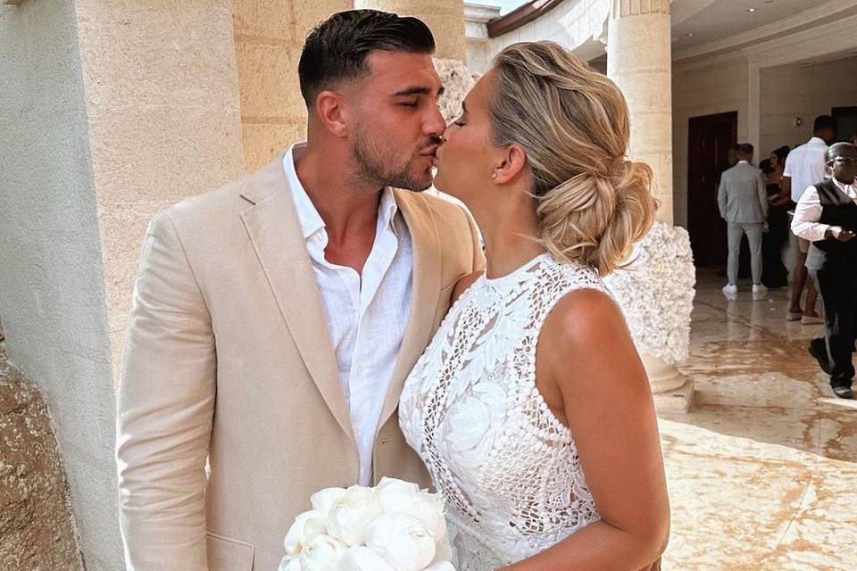 Molly-Mae wears a whte lace halterneck dress and holds a white bouquet as she kisses Tommy Fury wearing a beige suit