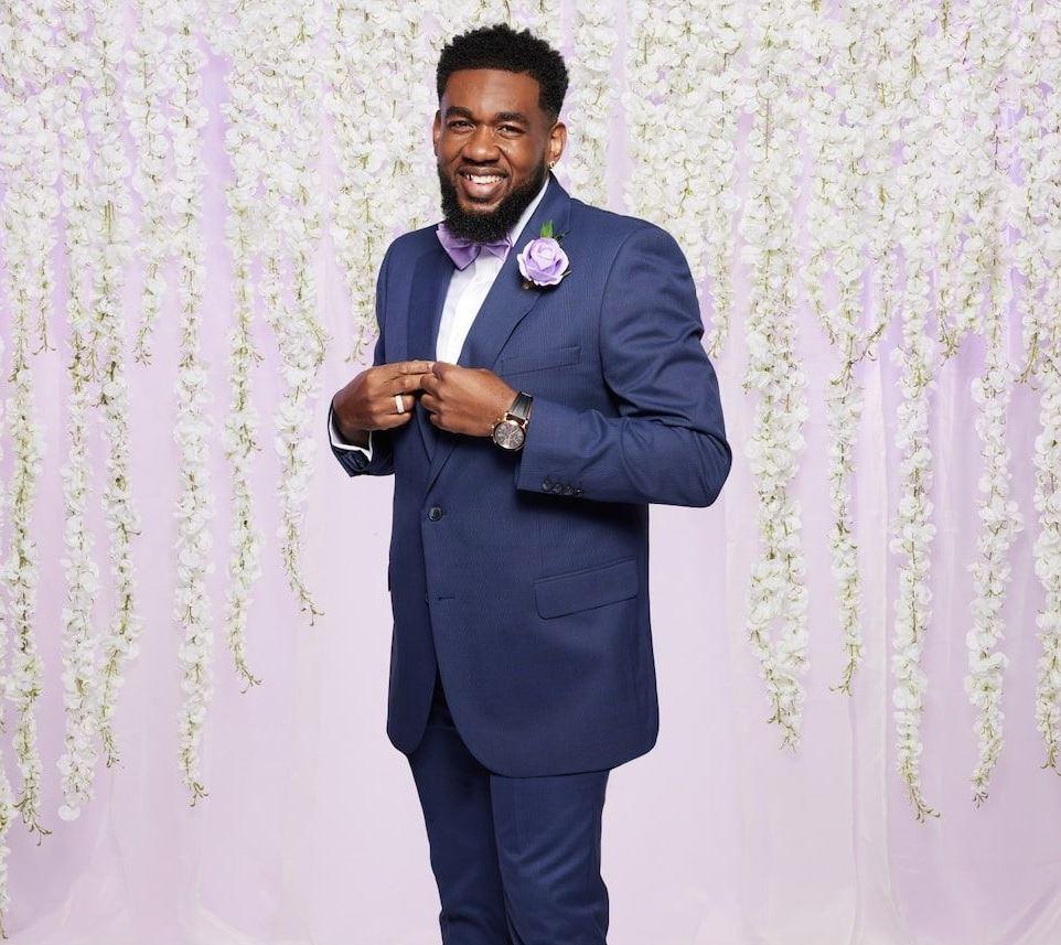 married at first sight groom terence wears a bright blue suit and purple bow tie in front of a lilac backdrop with white hanging flowers