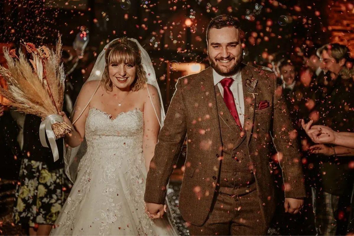 a bride with a rustic wedding bouqte and her husband in a brown tweed wedding suit leaving their ceremony at night with confetti around them