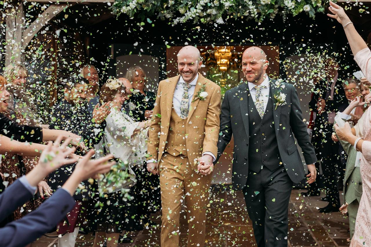 two grooms walking hand in hand out of a church into a stream of confetti at an LGBTQ+ wedding