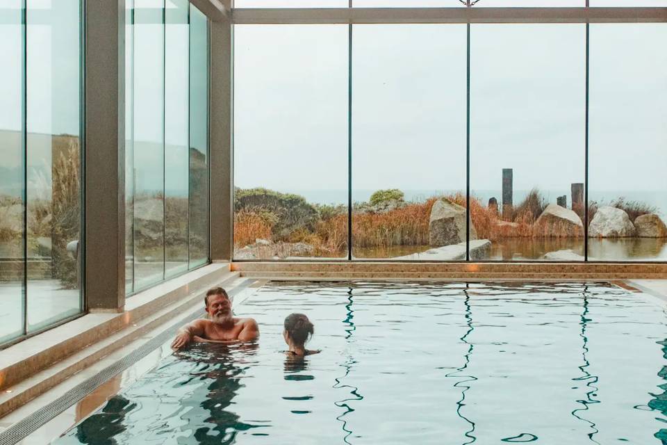 A man and woman in the indoor pool at a couples spa break in Cornwall enjoying unspoilt views of the ocean outside the windows