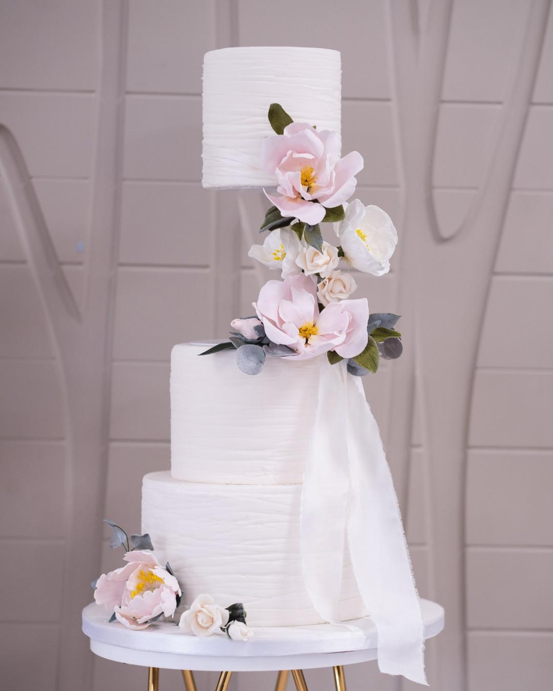 A simple three tier white wedding cake with sugar peonies on a gold cake stand.