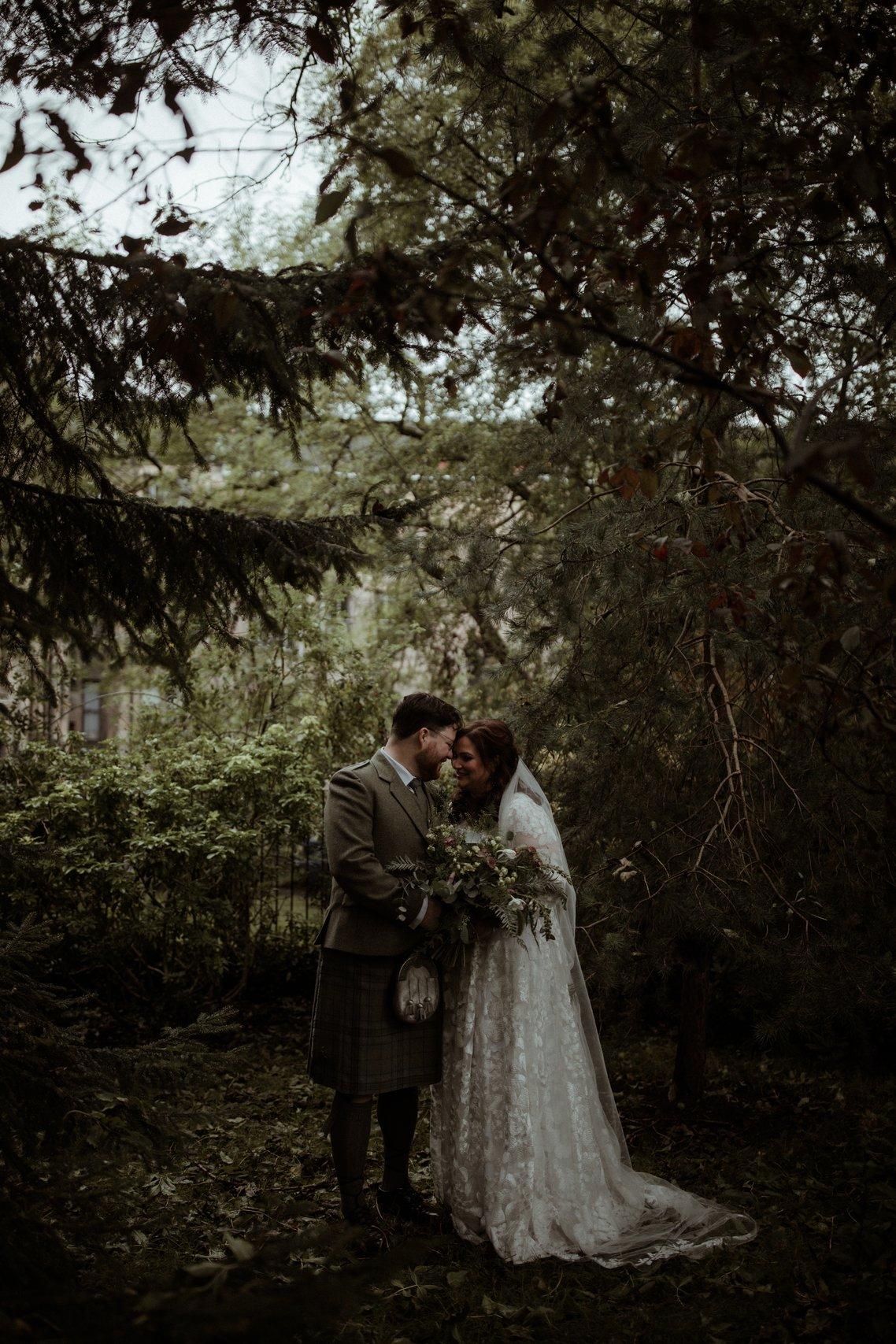 White newlywed couple standing at the bottom of a green garden smiling and touching foreheads with the kilt wearing a light green tweed suit and a kilt and the bride wearing an embroidered long sleeved white wedding dress with a low veil and green bouquet