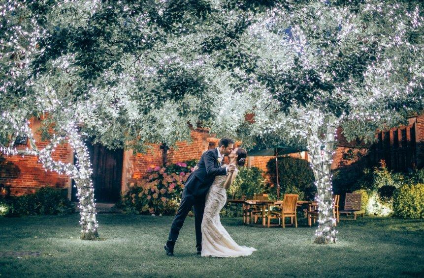 Couple kissing in wedding outfits under illuminated trees covered in fairy lights