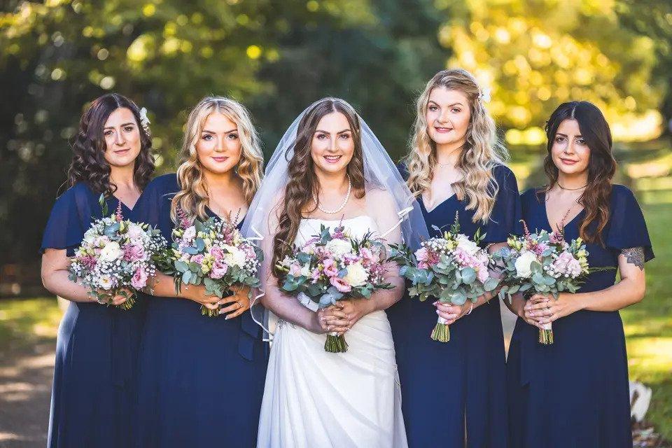 A traditional photo of a bride with two bridesmaids on either side of her smiling and holding bouquets wearing navy blue bridesmaid dresses