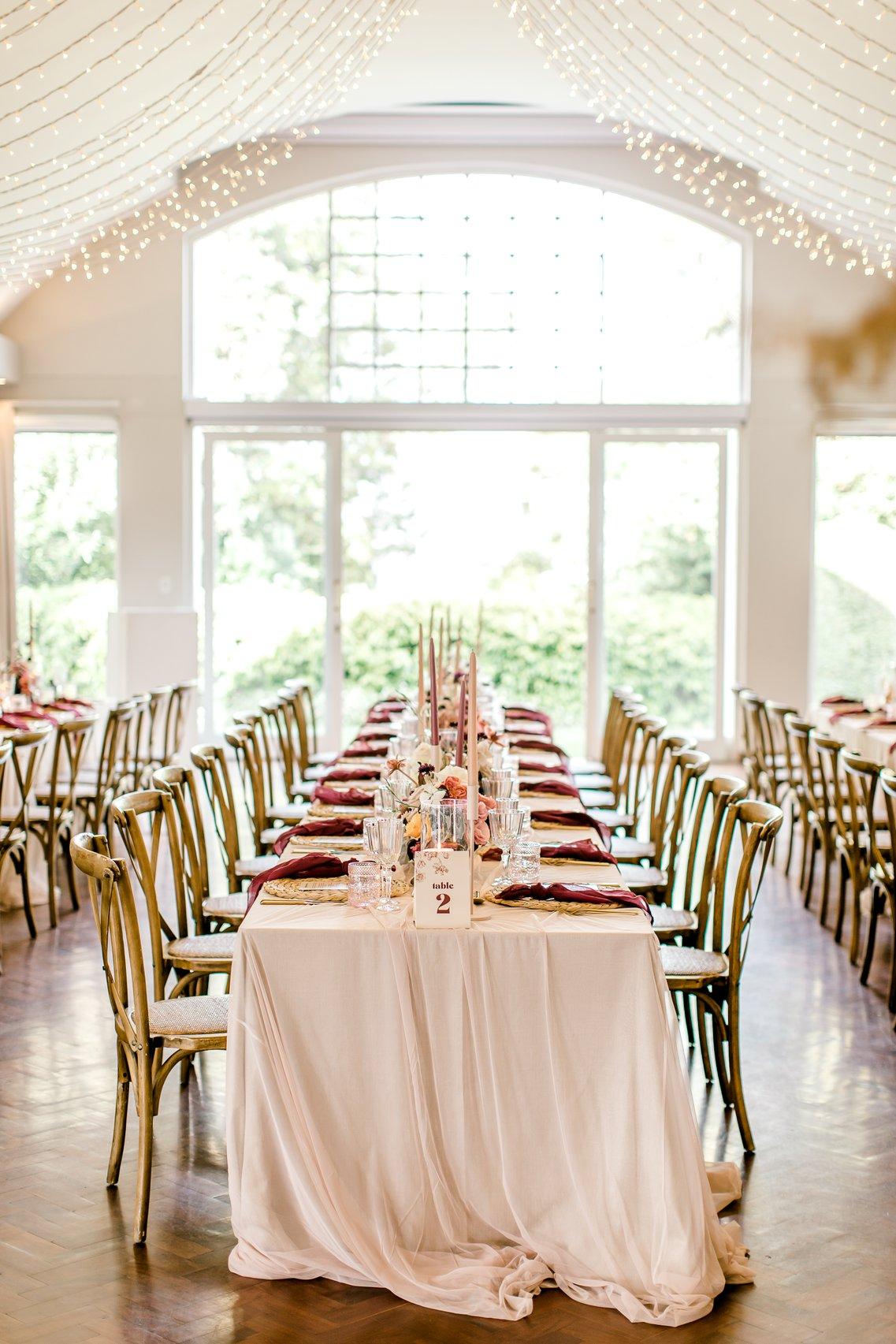 A front view of banquet style long tables and a glamorous wedding tablescape with white linens, rattan place mats and burgundy napkins