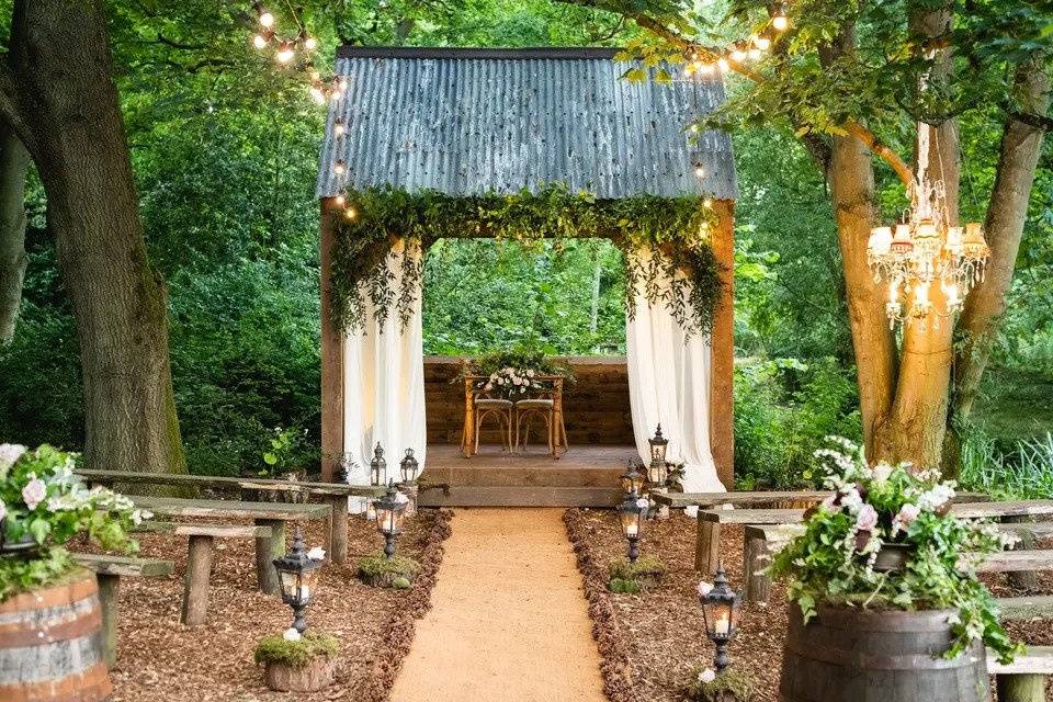 Wedding ceremony layout in a forest with wooden benches, an aisle lined by lampposts and two chairs placed under a wooden structure lined with greenery and curtains