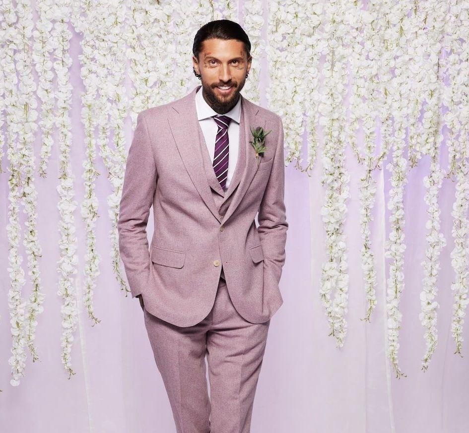 mafs uk groom wears a pastel pink suit and red tie as he stands with his hands in his pocket in front of a lilac backdrop with white hanging flowers