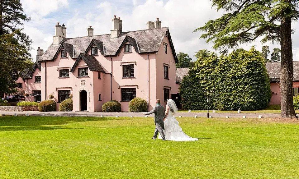 Bride and groom visible from the back as they walk through the grounds of their wedding venue with a pink coloured stately home wedding venue in the background