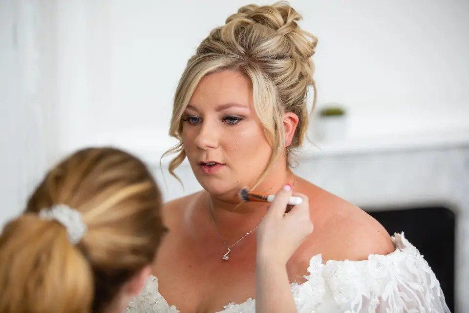 A seated bride with an elaborate updo has her makeup adjusted