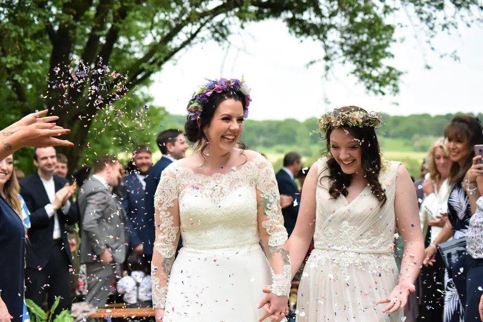 two brides wearing flower crowns walk outdoors while their guests throw confetti